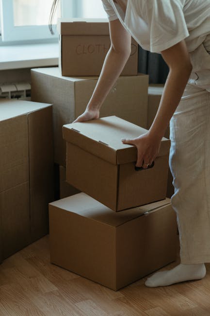 A person dressed in casual clothing is seen stacking and moving cardboard boxes inside a well-lit room with large windows. The boxes, made of sturdy brown cardboard, vary in size and are being carefully handled and arranged on a wooden floor, likely as part of a home relocation process. One box has the word 'CLOTHES' handwritten on it, indicating its contents. The individual is in the process of lifting a medium-sized box, preparing it for transport or placement, which is typical during packing and loading for a house move. In the background, additional boxes are visible, some placed on top of each other, suggesting a systematic approach to packing. This scene reflects the careful handling and organization involved in professional removals, such as those offered by Knightsbridge Removals, ensuring items are securely packed and efficiently loaded for safe furniture transport or home relocation. The environment appears tidy, with natural light illuminating the space, emphasizing attention to detail in the moving process.