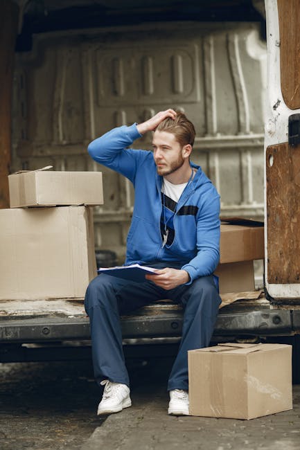 A young man with light brown hair, wearing a blue zip-up hoodie, navy trousers, and white sneakers, is sitting on the edge of the open back of a white moving van. He is holding a clipboard in his left hand and appears to be thinking or reviewing a list. Surrounding him are several cardboard boxes, some stacked and others on the ground, indicating a house removal or home relocation process. Inside the van, additional packing materials and boxes are visible, with the van’s interior featuring a metal lining and wooden panels. The scene is set outdoors on a paved surface, with the van positioned near a building or driveway. The lighting is natural, and the overall context suggests careful planning and organisation typical of furniture transport or packing during a residential move, with Knightsbridge Removals offering professional moving services. This visual accurately reflects aspects of loading and packing during removals.