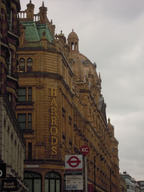 A close-up view of a historic sandstone building in Knightsbridge featuring elaborate architectural details, including domed roofs, decorative cornices, and ornate carvings. The facade displays the name 'HARRORDS' vertically along the corner of the building. In the foreground, a red and white London Underground bus stop sign is visible, indicating a transportation hub nearby. The setting appears to be in an urban area with a mix of classic and modern structures, suitable for home relocation or furniture transport services that involve careful packing, loading, and moving logistics as offered by Knightsbridge Removals.