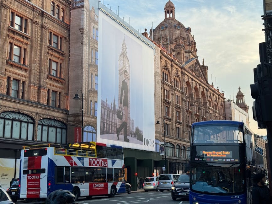 Photograph of a busy street scene in Knightsbridge with historic brick and stone buildings featuring ornate architectural details and large arched windows, under a partly cloudy sky with sunlight illuminating the facades. Prominently visible is a large billboard advertising Dior, mounted on a scaffolding structure attached to one of the buildings. In the foreground, a red and white double-decker bus operated by Tootbus London is stopped at the intersection, with the side displaying the company logo and route information. Next to it, a dark blue double-decker bus with an electronic destination sign indicating a route to Kensington is also visibly waiting or moving through the street. Several cars are lined up behind the buses, and a few pedestrians are walking along the sidewalk, some near the intersection. The scene captures London's architectural heritage alongside active urban transport, consistent with relocation and moving service logistics in an iconic central London setting, highlighting the environment for house removals, furniture transport, and packing and moving activities in accordance with Knightsbridge Removals.