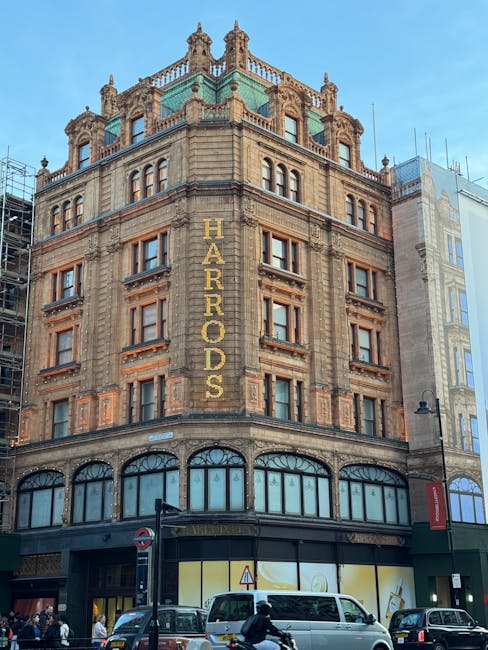 Photograph of the historic Harrods building in Knightsbridge, featuring its ornate stone façade with multiple levels of arched windows and decorative architectural details. The building displays a large vertical sign spelling 'HARRADS' in gold letters on the corner, and the lower ground floor has large glass display windows. In front of the building, the street scene includes a white delivery van positioned near the curb, pedestrians walking on the pavement, and several other vehicles including cars and a motorcycle. The scene is set during daylight with a clear blue sky, and nearby buildings can be seen, indicating an urban environment. This image reflects a typical location for house removals or furniture transport activities associated with professional moving services like Knightsbridge Removals, capturing the setting where coordination and logistics for house moves are undertaken.