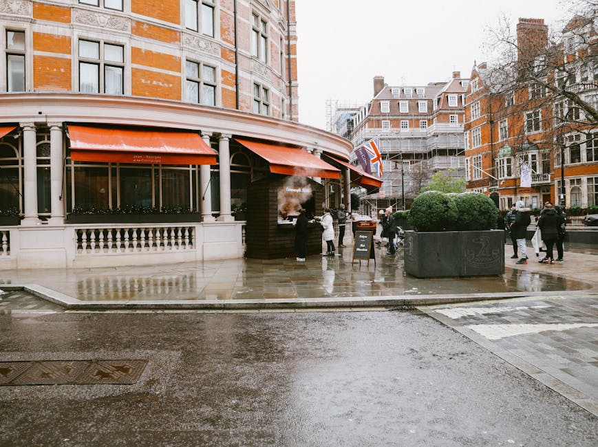 Photograph of a busy street scene in Knightsbridge with historic brick and stone buildings featuring ornate architectural details and large arched windows, under a partly cloudy sky with sunlight illuminating the facades. Prominently visible is a large billboard advertising Dior, mounted on a scaffolding structure attached to one of the buildings. In the foreground, a red and white double-decker bus operated by Tootbus London is stopped at the intersection, with the side displaying the company logo and route information. Next to it, a dark blue double-decker bus with an electronic destination sign indicating a route to Kensington is also visibly waiting or moving through the street. Several cars are lined up behind the buses, and a few pedestrians are walking along the sidewalk, some near the intersection. The scene captures London's architectural heritage alongside active urban transport, consistent with relocation and moving service logistics in an iconic central London setting, highlighting the environment for house removals, furniture transport, and packing and moving activities in accordance with Knightsbridge Removals.