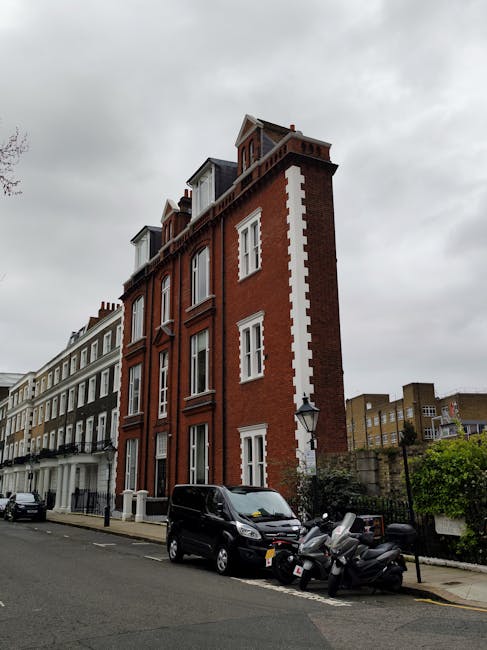 A multi-story red brick building with white window frames and decorative elements, situated on a city street under a cloudy sky. In front of the building, a black van is parked on the street, alongside several parked motorcycles, with protective plastic and fabric wrapping visible on some furniture pieces. The scene suggests recent or ongoing home relocation activities, with the vehicle and motorcycles positioned close to the building's entrance, which is not directly visible. The surrounding area features other similar residential buildings, a lamppost, and some greenery, reflecting an urban environment typical for house removals and furniture transport in Knightsbridge. This setting aligns with professional removals services, such as those provided by Knightsbridge Removals, supporting packing, loading, and transportation processes involved in house moves and furniture relocations.