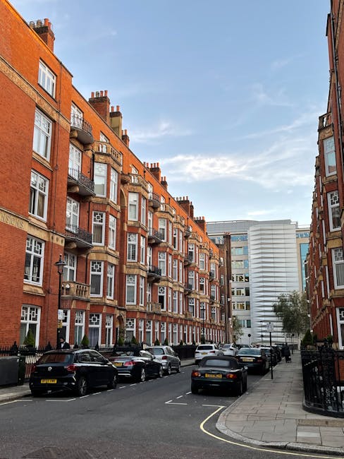 A street scene in a residential area with a row of multi-storey red-brick buildings featuring white-framed windows and small balconies, some with decorative railings. Several parked cars line the street, including black and silver vehicles, with a UK-style license plate visible on one of the cars. The pavement runs alongside the buildings, leading to a black fence and gated entrance. In the background, a modern white multi-storey building contrasts with the older architecture. The sky is partly cloudy with patches of blue, and the overall scene suggests an urban environment suitable for house removals and furniture transport, with no activity currently visible in the loading area. The image supports content related to home relocation, packing, and moving logistics, such as those provided by Knightsbridge Removals.