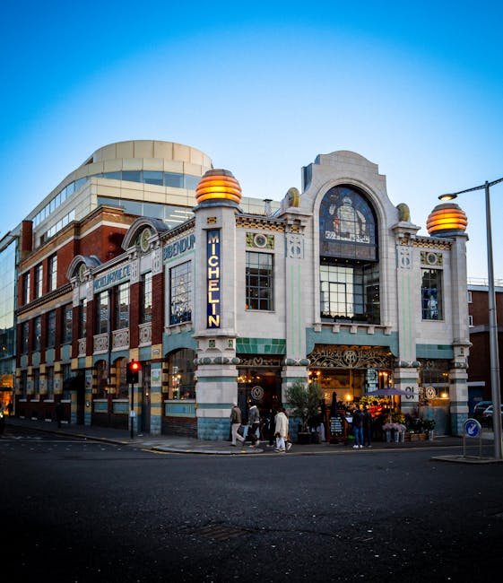 Exterior view of a multi-story building at the corner of a city street during daytime, with large windows and decorative architectural features. The building has signage indicating it is the Michelin House, with a vertical neon sign reading 'MICHELIN' attached to a column at the street corner. In front of the building, there is a pedestrian crossing with a traffic light showing red, and several people are walking along the pavement or waiting to cross. The street is lined with lamp posts, and the scene depicts a typical urban environment. This setting could be part of a property where house removals or relocation services by Knightsbridge Removals are conducted, involving careful planning of furniture transport and packing during a home relocation process.