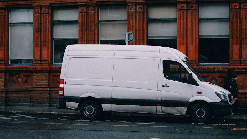 A white commercial van parked parallel along a street in front of a historic red brick building with decorative stone window surrounds. The van's side door is closed, and it appears to be used for furniture transport during a home relocation. In the background, two individuals are visible, one standing near the building's entrance and the other walking past, suggesting a moving or packing process. The scene is captured in daylight with clear weather, and the van is positioned close to the curb, ready for loading or unloading of cardboard boxes, plastic-wrapped furniture, or other packing materials typical of professional removals. Knightsbridge Removals employs such vehicles for efficient home relocation services, especially when navigating narrow access points in urban environments like Sloane Street, as discussed in the page about tips for narrow access moving from Knightsbridge. Furniture and packing materials are likely inside the van or on the pavement, awaiting transfer during the loading process.