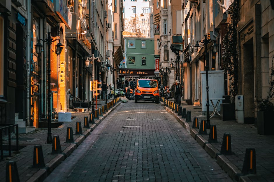 A narrow urban street in Knightsbridge with tall, closely spaced buildings on both sides, featuring shopfronts and residential entrances. The cobblestone pavement is flanked by yellow and black bollards, guiding vehicle and pedestrian movement through the constrained space. An orange van is parked at the end of the street, near a building with a green facade, while a few people are visible in the background, some standing and others moving. The scene is lit by natural daylight, casting shadows on the buildings and street, reflecting typical conditions for house removals or furniture transport through tight access routes. This setting illustrates the challenges of home relocation in narrow streets and highlights the importance of careful planning by Knightsbridge Removals for loading and unloading furniture and moving boxes in confined environments such as this, with attention to street layout and spatial limitations.