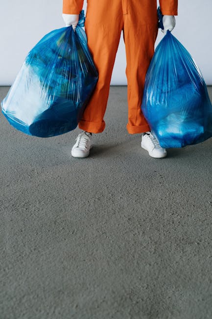 A person dressed in orange overalls and white shoes is standing inside a room with a plain light-colored wall and a gray carpeted floor, holding two large blue plastic garbage bags filled with waste,