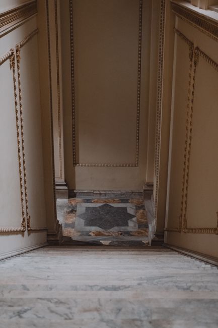 A view from the top of a staircase looking down into a narrow residential corridor, with the staircase featuring white marble steps. On either side of the staircase, there are cream-colored wooden walls decorated with ornate gold trim and beaded moldings, indicating a period-style interior. At the bottom of the stairs, the corridor floor is covered with patterned tile, and a small section of a wooden doorframe is visible at the far end. In the context of house removals or furniture transport, Knightsbridge Removals may conduct moving services involving narrow staircases like this, requiring careful planning and precise furniture handling within confined spaces.