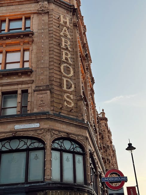 Photograph of a historic multi-storey building in Knightsbridge, London, with the word 'THE ARCADE' displayed vertically in large, illuminated letters on its ornate brick and stone façade. The building features decorative architectural details, including arched windows with intricate framing on the ground floor. Adjacent to the building, there is a classic street lamp and a London Underground station sign indicating proximity to public transport. The sky is clear with minimal clouds, and the overall scene emphasizes the exterior of the building in a city environment. This setting relates to house removals and furniture transport, as it suggests an urban area where professional movers, like Knightsbridge Removals, often assist with building clearance, packing, and moving services to facilitate home relocation or commercial moves within or around Knightsbridge.