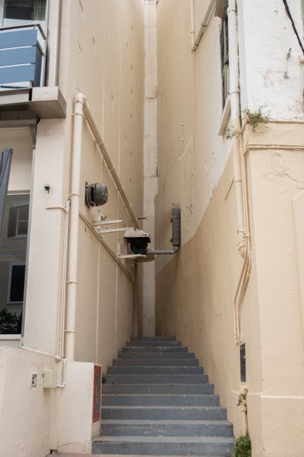 A view from the top of a staircase looking down into a narrow residential corridor, with the staircase featuring white marble steps. On either side of the staircase, there are cream-colored wooden walls decorated with ornate gold trim and beaded moldings, indicating a period-style interior. At the bottom of the stairs, the corridor floor is covered with patterned tile, and a small section of a wooden doorframe is visible at the far end. In the context of house removals or furniture transport, Knightsbridge Removals may conduct moving services involving narrow staircases like this, requiring careful planning and precise furniture handling within confined spaces.